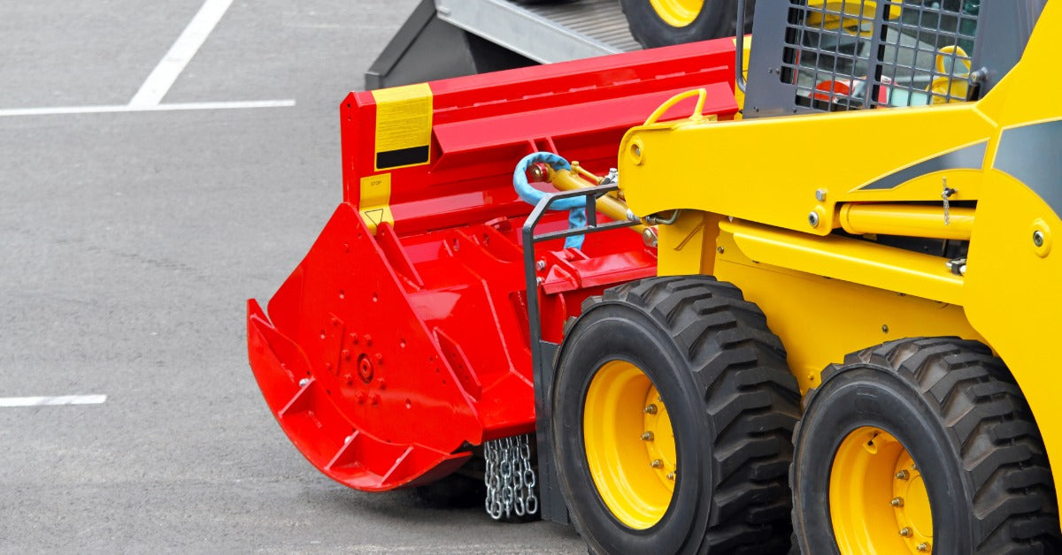 A close-up of a yellow skid steer with a bright red attachment parked on a section of concrete with painted lines.