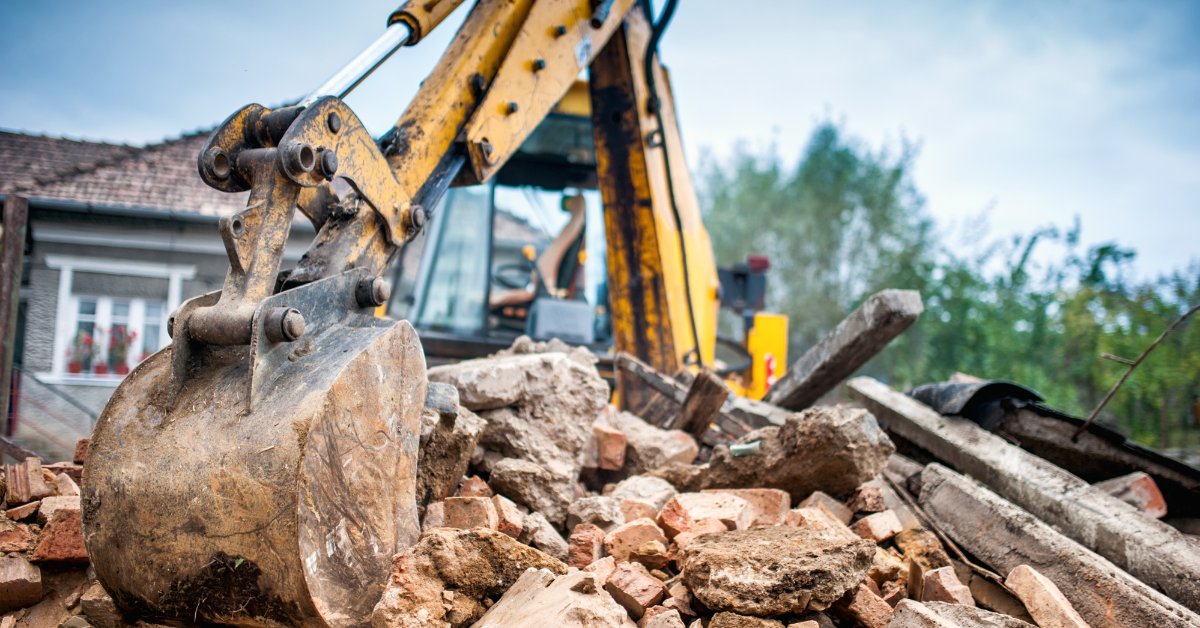 A yellow excavator bucket resting on a pile of broken bricks, concrete, and construction debris outdoors.