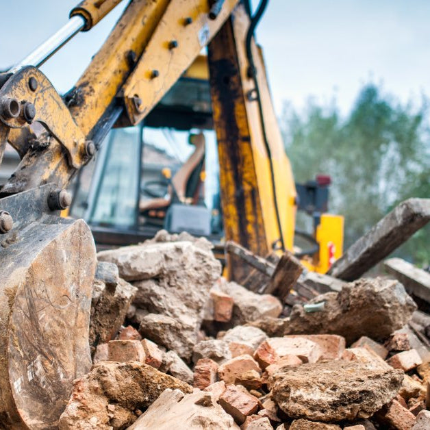 A yellow excavator bucket resting on a pile of broken bricks, concrete, and construction debris outdoors.