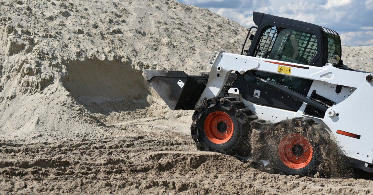 A white skid steer loader with a front bucket moving through a large mound of sand under a cloudy sky.