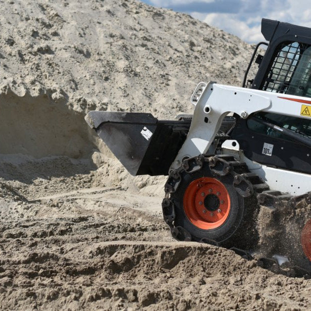 A white skid steer loader with a front bucket moving through a large mound of sand under a cloudy sky.