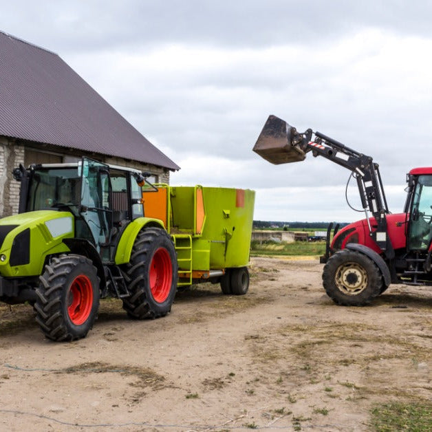 Two tractors operating on a farm. One tractor has a bucket attachment and the other is pulling a trailer with a feed distributor.