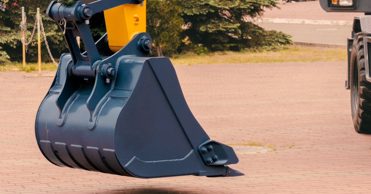 A close-up of a gray metal bucket attached to the yellow arm of an excavator. The machine is parked on a brick surface.