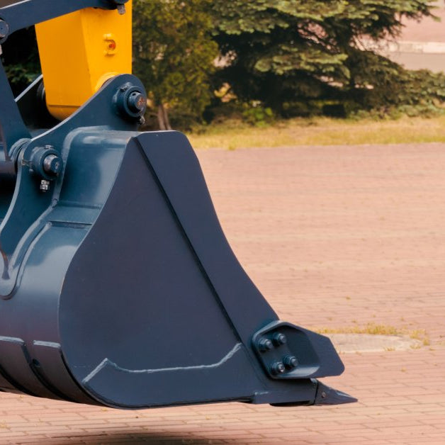 A close-up of a gray metal bucket attached to the yellow arm of an excavator. The machine is parked on a brick surface.