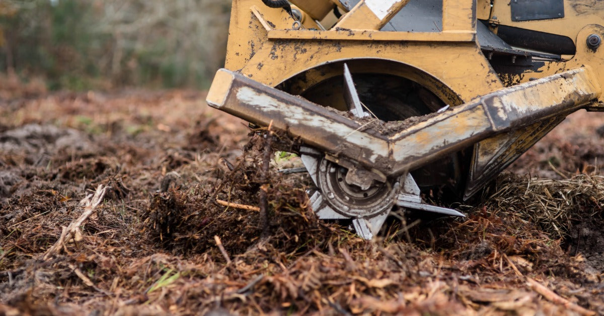 A close-up of a yellow brush cutter attachment resting on a pile of shredded debris in the middle of a wooded area.