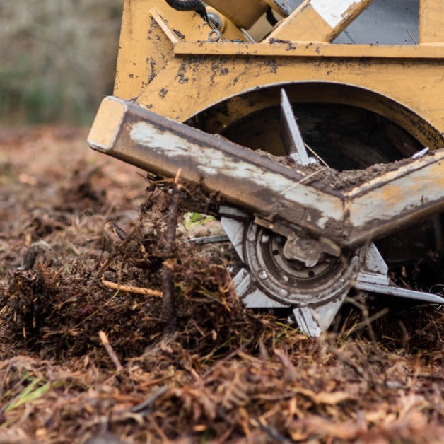 A close-up of a yellow brush cutter attachment resting on a pile of shredded debris in the middle of a wooded area.