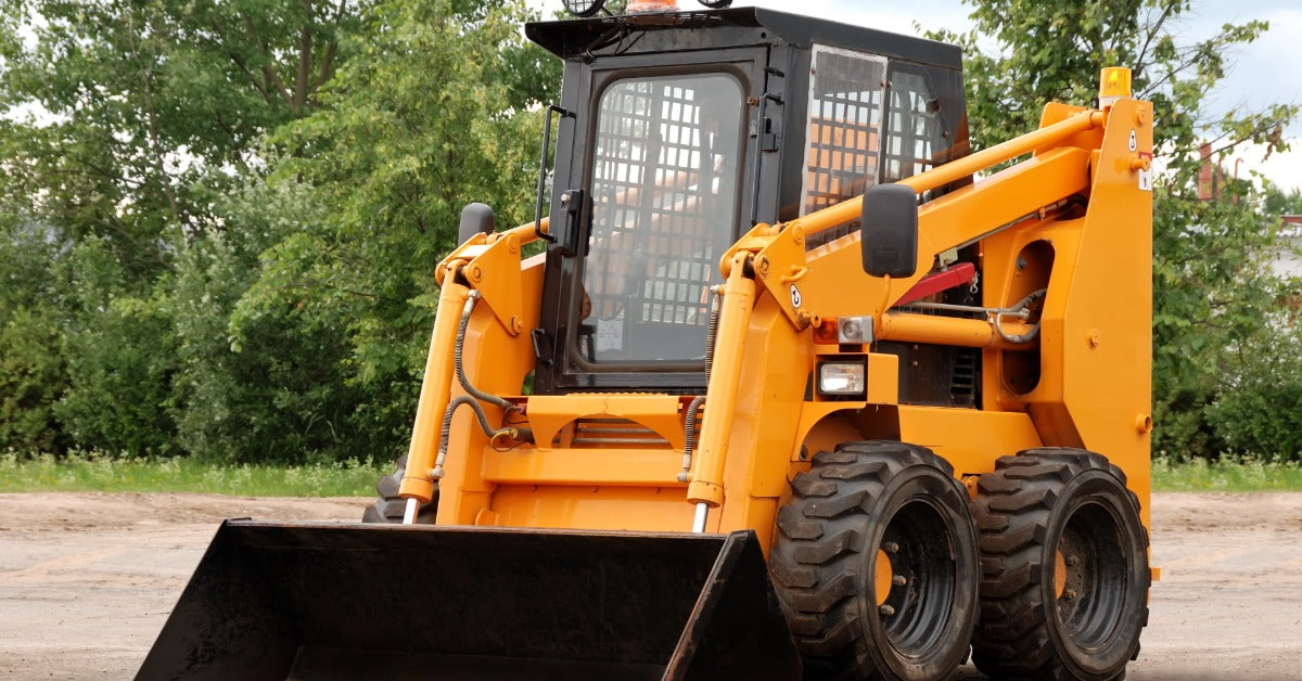A close-up of a yellow skid steer with a bucket attachment. The machine is parked on a space of compacted dirt.