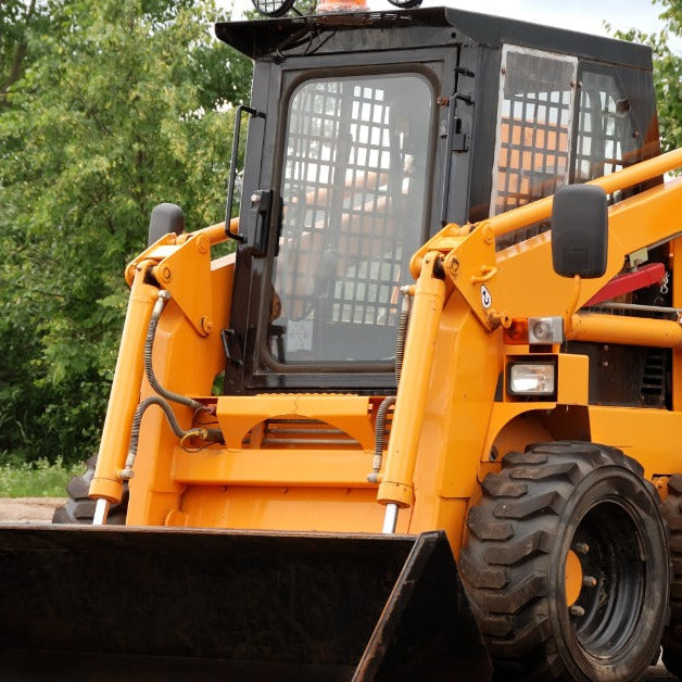A close-up of a yellow skid steer with a bucket attachment. The machine is parked on a space of compacted dirt.